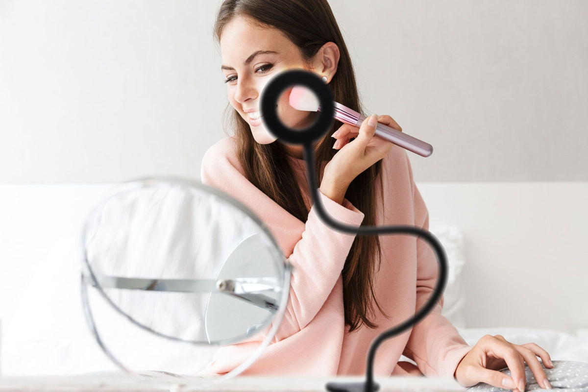 A woman smiles on her bed while applying makeup, using the Rio Beauty Station LED Ring Light for soft illumination. In front of her are a round mirror and a black flexible phone holder, highlighting her long brown hair and light pink top.