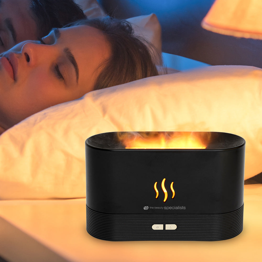 A woman sleeps peacefully beside a nightstand holding the Rio ALTA Aroma Diffuser, Humidifier and Night-light, which glows with a warm, flame-like light and features wavy steam symbols and control buttons on its front.