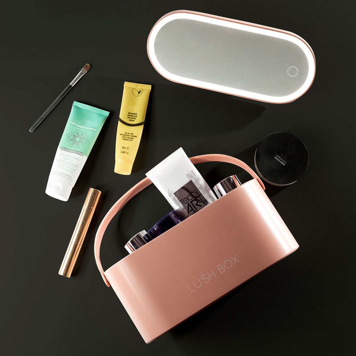 A pink Lush Box Vanity Case by Rio sits on a dark surface, surrounded by skincare tubes, a makeup brush, a mascara tube, and an oval mirror with a button.