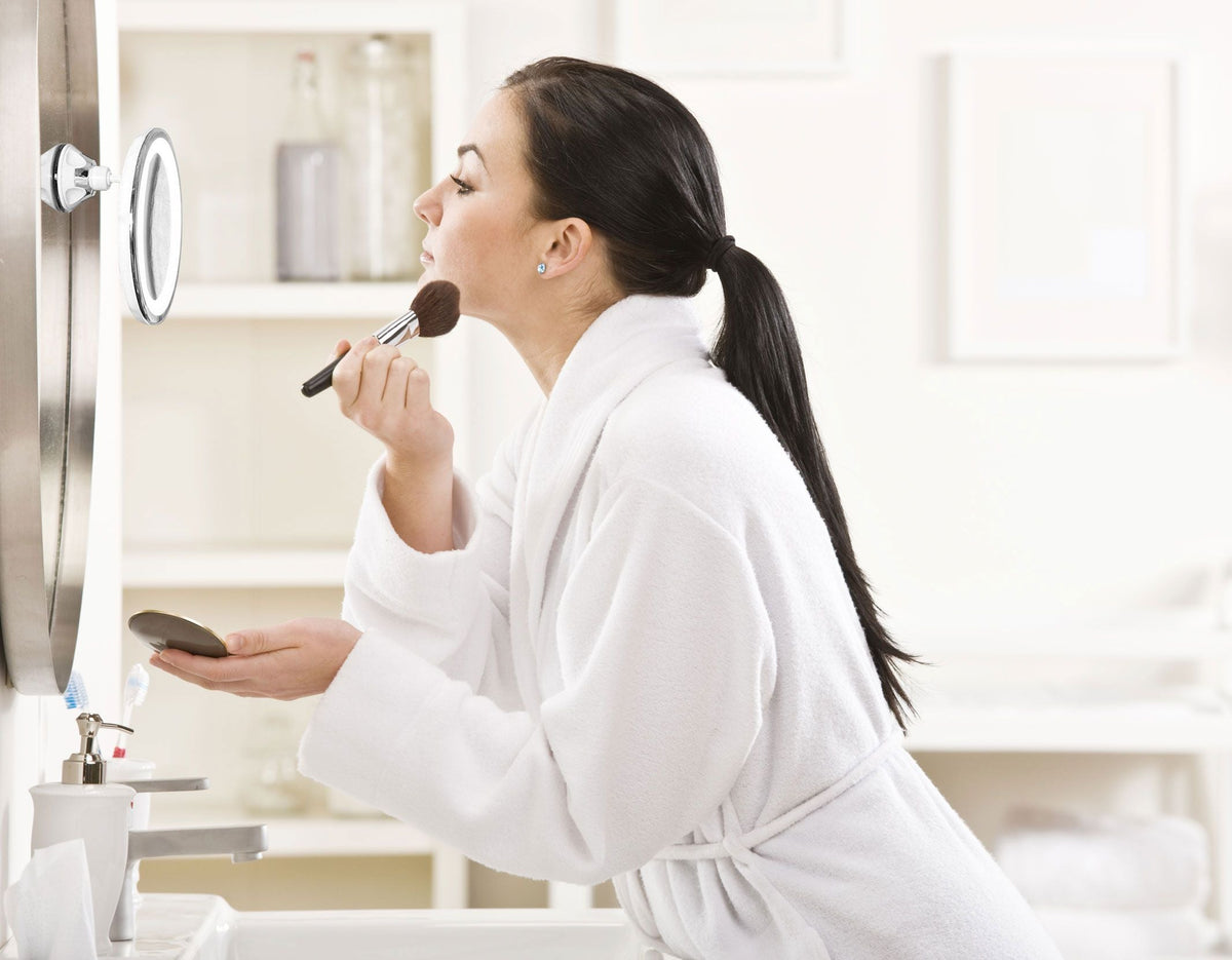 lady using Multi use LED illuminated make up mirror with suction cup base attached to larger wall mounted mirror in bathroom applying make up 